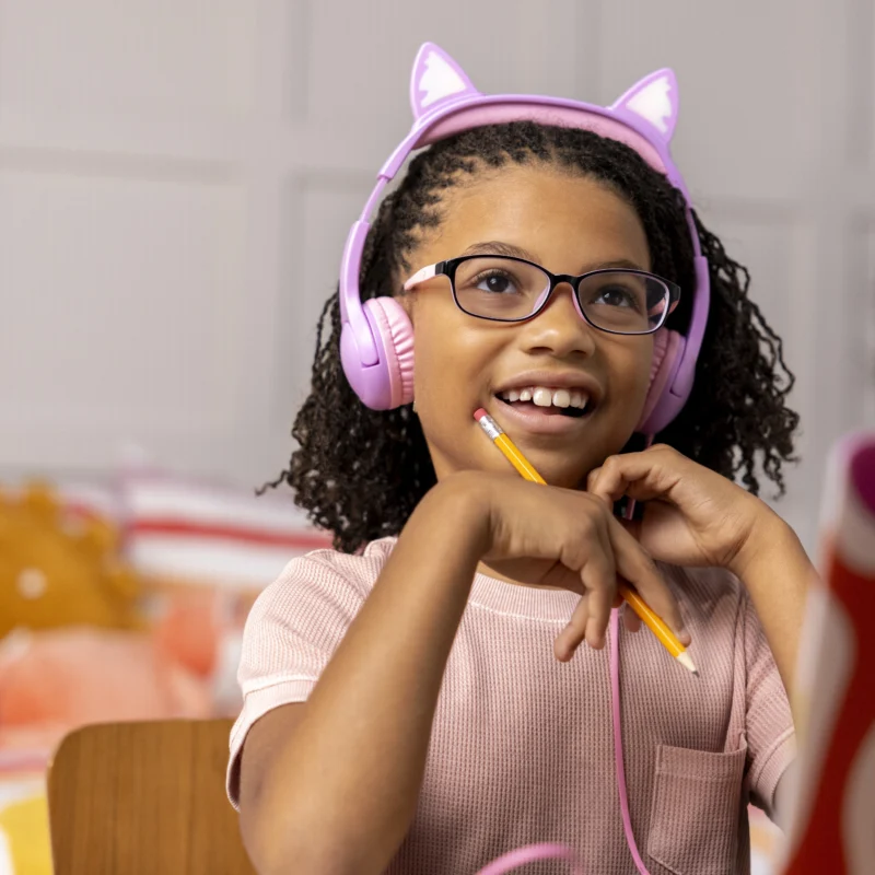 A smiling girl with curly hair and glasses wears pink cat-ear headphones, holding a pencil while using an online learning platform at her desk with open books and a laptop.