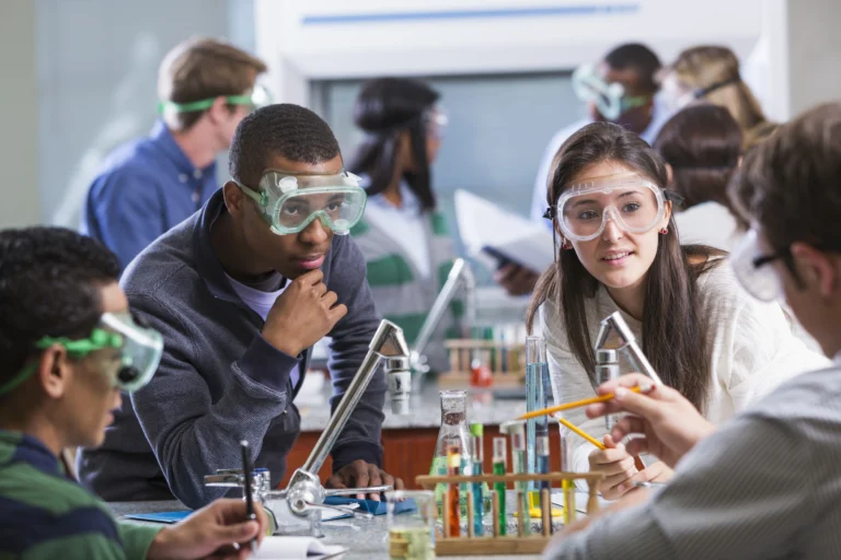 A group of high school students wearing safety goggles work together in a science lab, discussing an experiment with test tubes and lab equipment, while using an online learning platform to enhance their understanding.