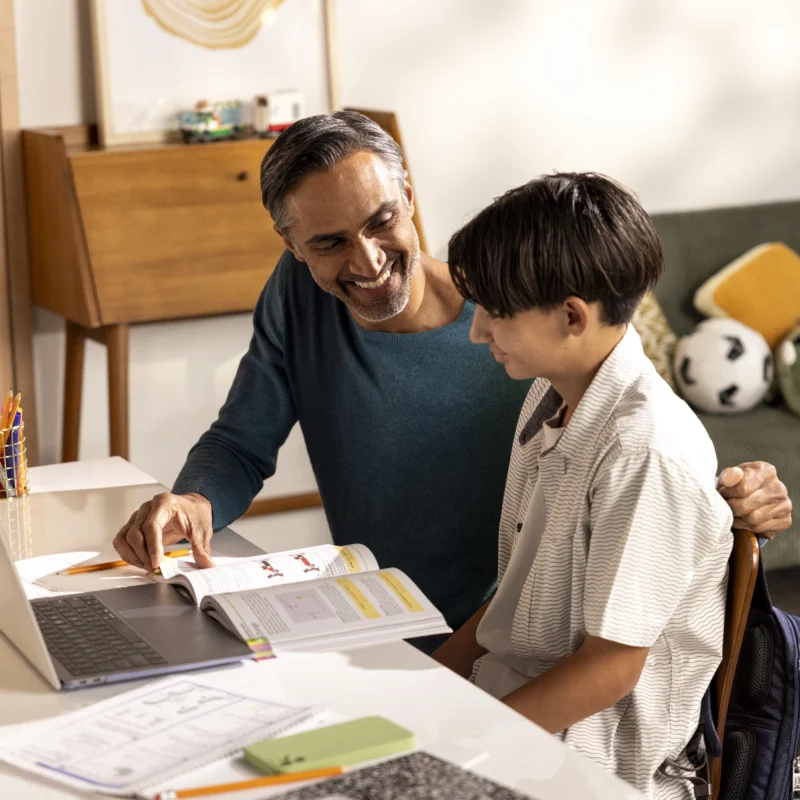 An adult and a child sit at a desk with a laptop and books, smiling and studying together in a well-lit room. A couch with soccer balls and a guitar is visible in the background, highlighting the fun of an online learning platform.