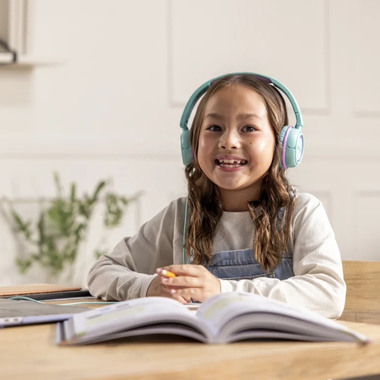 A smiling young girl wearing teal headphones sits at a table with an open book and notebooks, appearing to study or do homework in a bright, cozy room—perfectly immersed in her online learning platform.