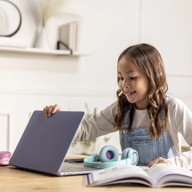 A young girl sits at a wooden table, smiling at a laptop from an online learning platform. Open books and headphones are in front of her, in a bright, cozy room with shelves in the background.
