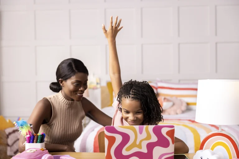 A smiling woman sits beside a young girl with raised hand as she uses an online learning platform on a colorful laptop at a desk in a bedroom decorated with bright patterns and rainbows.