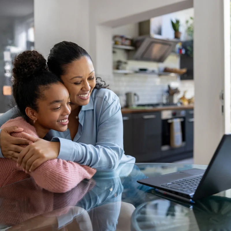 A woman hugs a smiling girl from behind as they sit at a glass table with an open laptop, using an online learning platform in a bright, modern kitchen.