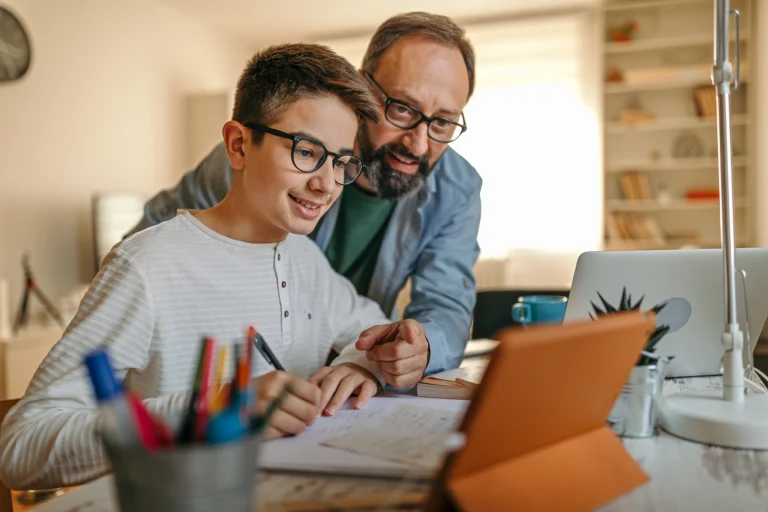 A smiling boy wearing glasses writes in a notebook while a man leans over his shoulder, both looking at a tablet on a desk with school supplies and a laptop, exploring an online learning platform in a cozy, well-lit room.
