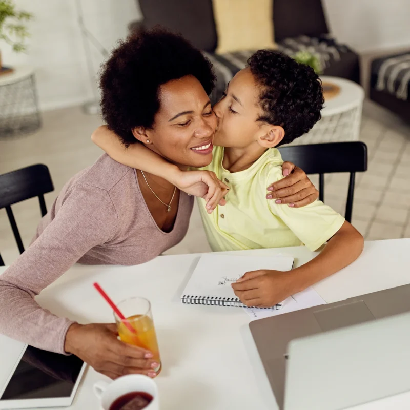 A smiling woman sits at a table with a notebook and laptop, exploring digital learning solutions for schools, as a young boy hugs and kisses her cheek. A glass of orange juice and a cup are also on the table.