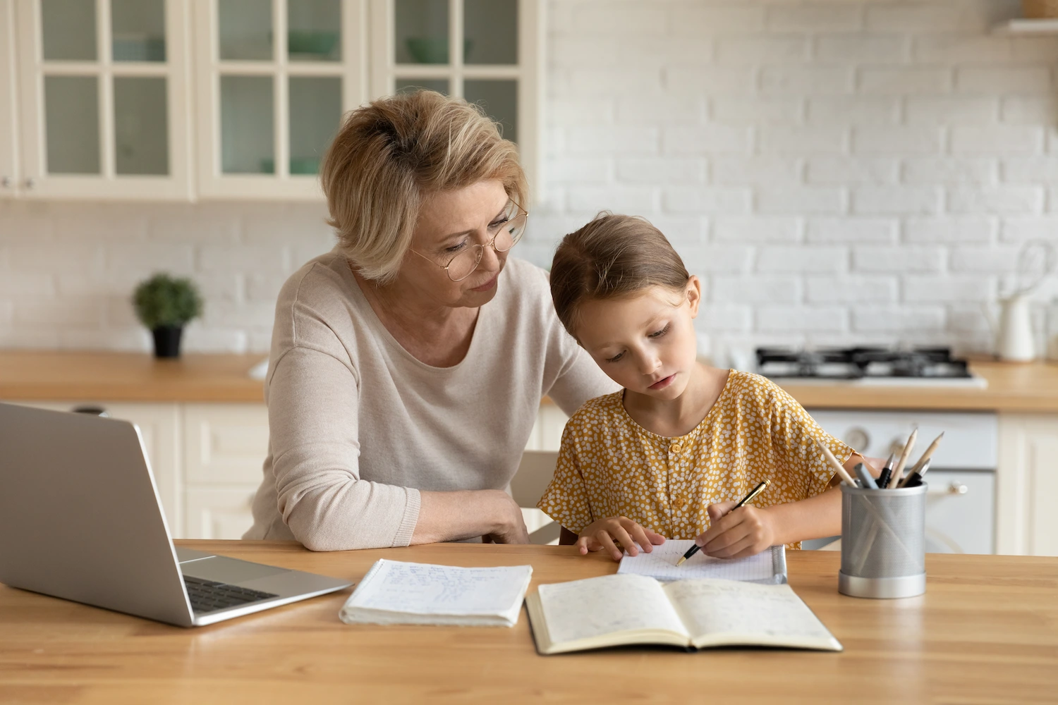 An older woman and a young girl sit at a kitchen table. The woman watches as the girl writes in a notebook, creating an atmosphere reminiscent of a Learning Hub. A laptop, papers, and a pencil holder are on the table, with the kitchen in the background.