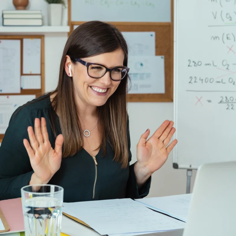 A woman with glasses smiles and raises her hands while sitting at a desk, looking at a laptop from an online learning platform. She wears earbuds, and a whiteboard with math equations is visible behind her in a bright home office.