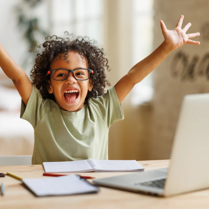 A smiling child wearing glasses sits at a desk with a laptop, notebooks, and colored pencils, raising their arms in excitement while celebrating an achievement on an online learning platform at home.