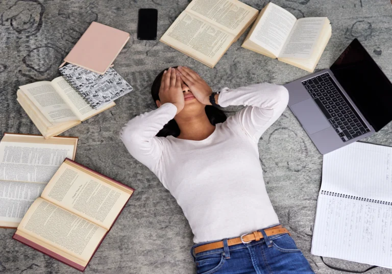 A person lies on the floor covering their face with their hands, surrounded by open books, a laptop, a phone, and notebooks—appearing stressed or overwhelmed while studying on an online learning platform.