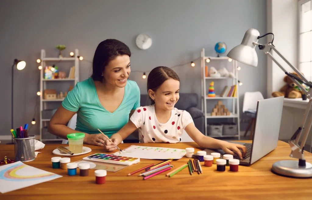 A woman and a girl sit at a desk with art supplies, painting and smiling while looking at a laptop in a brightly lit room with shelves and decorations in the background, as they explore creative ways to support measures of academic progress.