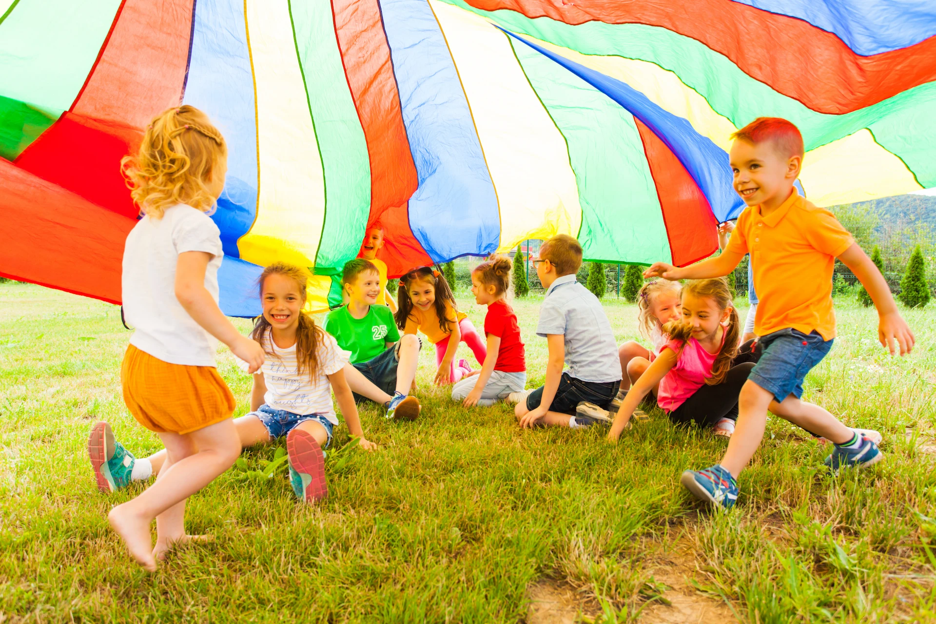 A group of young children play together outside, sitting and running under a large, colorful parachute spread above them on the grass, smiling and laughing in the sunlight as they experience Learning Through Play.