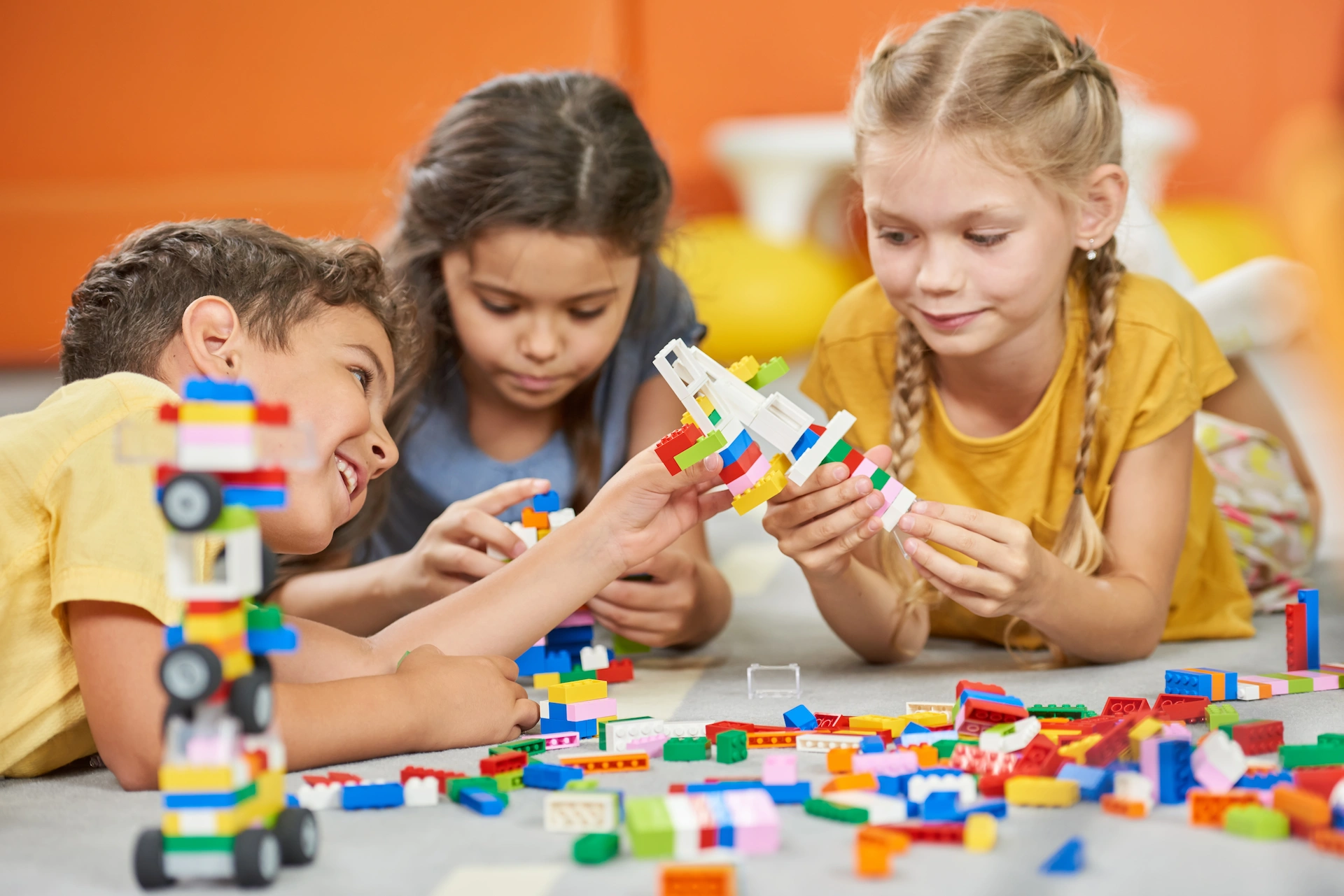 Three young children playing together on the floor with colorful plastic building blocks, smiling and focused as they construct various creations.