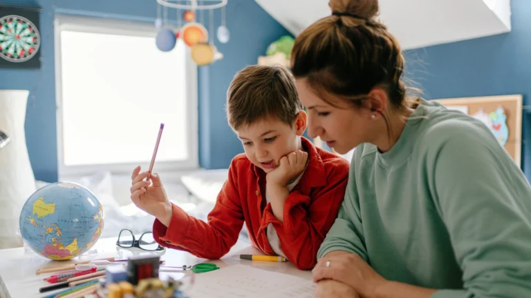 A woman and a young boy sit at a table, working on homework together. The boy holds a pencil while the woman looks at the paper, helping him stay on track with their easy homeschool schedules. A globe, colored markers, and glasses are on the table. The room is brightly lit.