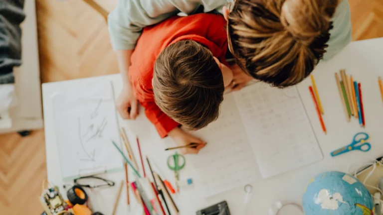 A child and an adult sit at a table, working together on schoolwork. Colored pencils, paper, and a globe are spread out on the table as they find your family’s flow in learning together.