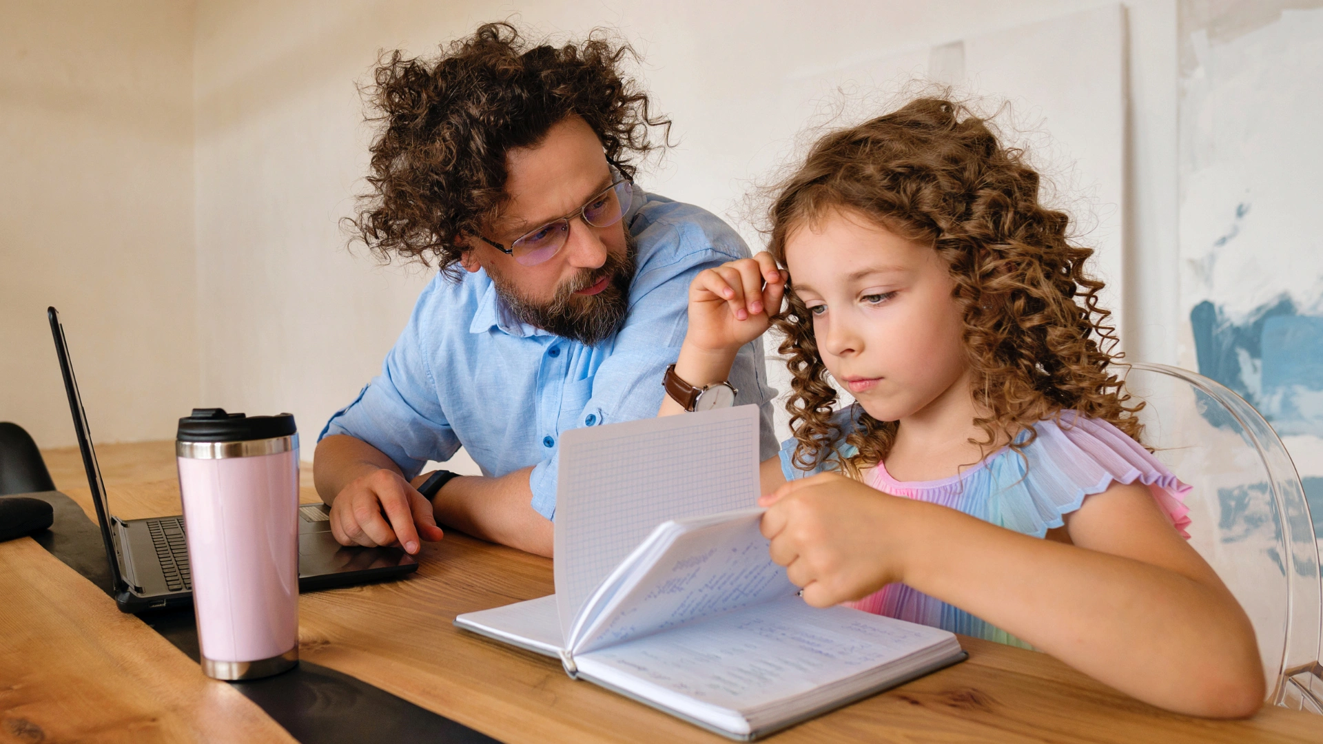 During their Morning Homeschool Routine, a man with curly hair and glasses helps a young girl with curly hair as she reads from a notebook at a wooden table, with a laptop and pink travel mug beside them.