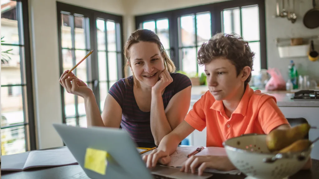 A woman and a boy sit at a table, smiling and looking at a laptop as they enjoy some gentle homeschool planning together. The boy wears an orange shirt and types, while the woman holds a pencil. Sunlight streams through large windows behind them.