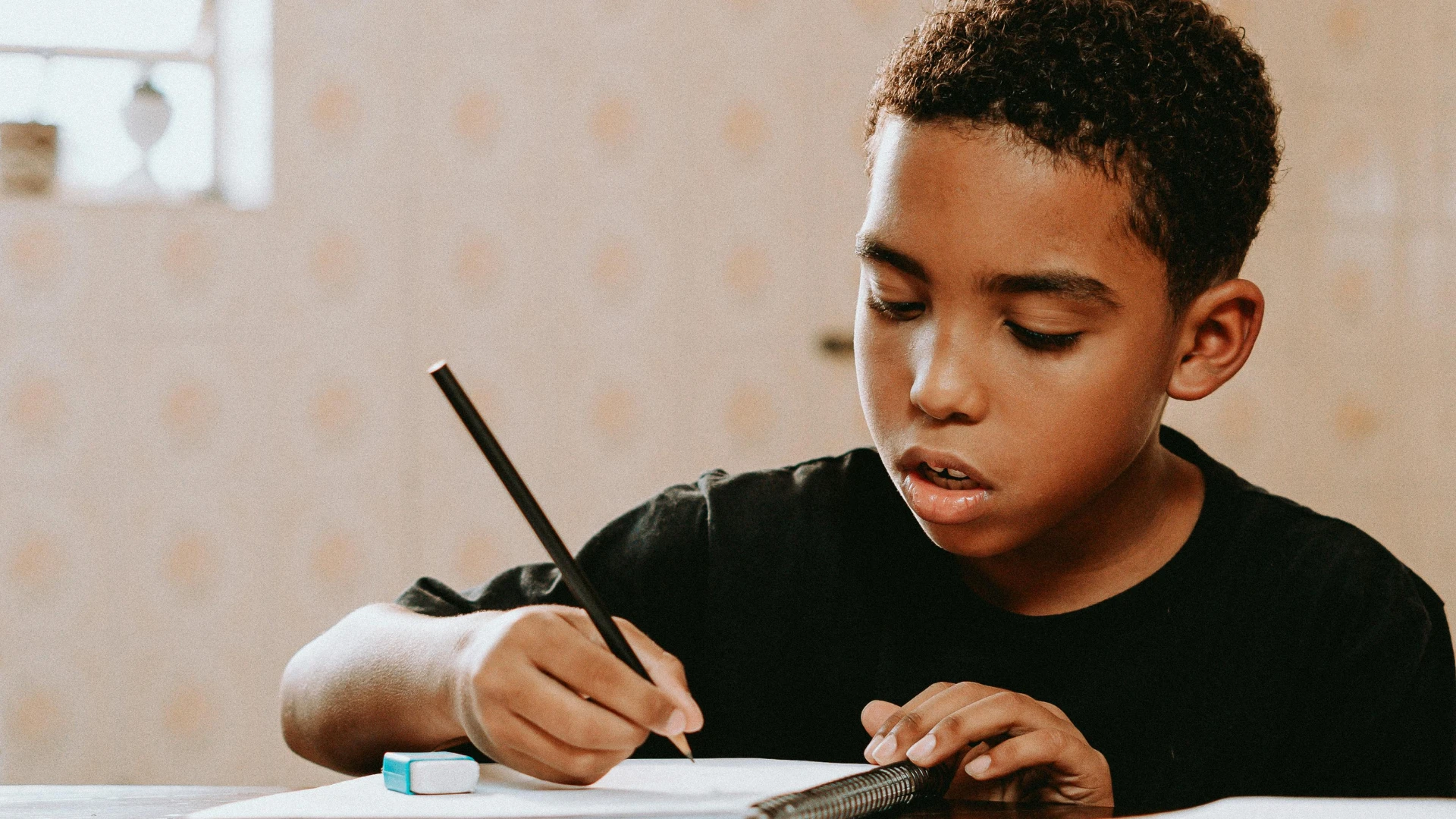 A young boy with short curly hair, wearing a black shirt, sits at a table and writes on paper with a pencil. An eraser and pencil case are on the table. He appears focused on his work.