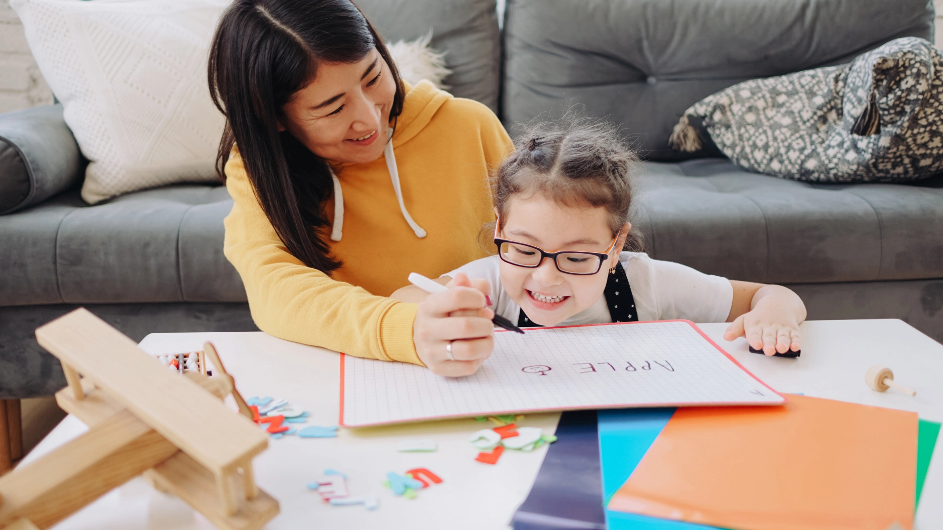 A woman helps a young girl with glasses write “APPLE” in a notebook at a table covered with colorful folders, letter tiles, and a wooden toy, while sitting in a cozy living room.