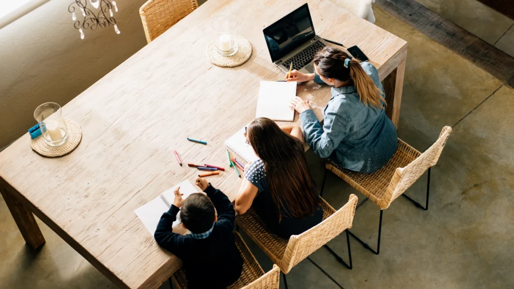 Three people sit at a wooden table with notebooks, colored pens, and a laptop, viewed from above. They appear to be working or studying together in a well-lit room, reflecting the collaborative spirit often found in a homeschool community.
