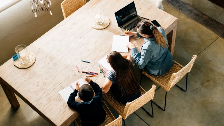 Three people sit at a wooden table with notebooks, colored pens, and a laptop, viewed from above. They appear to be working or studying together in a well-lit room, reflecting the collaborative spirit often found in a homeschool community.