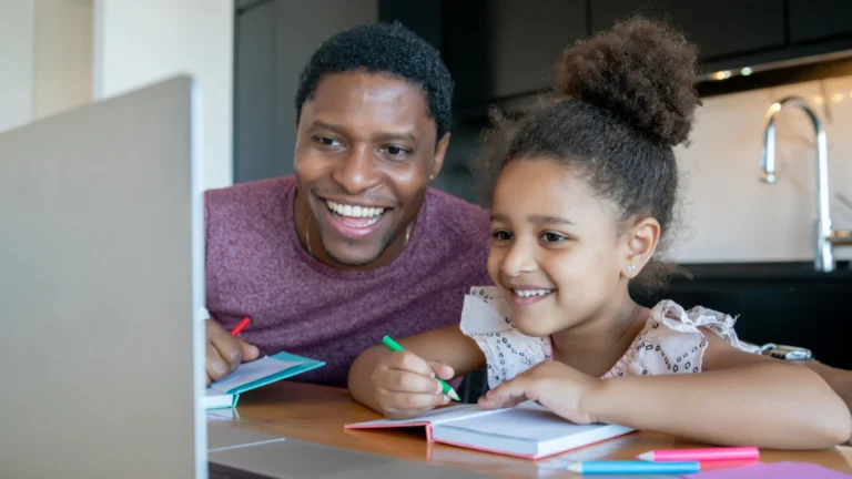 A man and a young girl smile while sitting at a table, enjoying some of the homeschool co-op benefits as they write in notebooks with colored pencils and look at a computer screen.