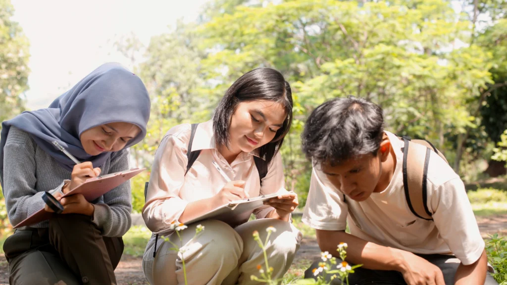 Three young people sit outdoors, closely examining plants and taking notes on clipboards. Sunlight filters through trees in the background, creating a bright, natural setting—an ideal scene for gathering Nature Exploration Tips.