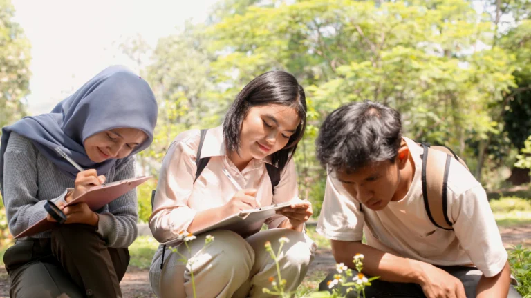 Three young people sit outdoors, closely examining plants and taking notes on clipboards. Sunlight filters through trees in the background, creating a bright, natural setting—an ideal scene for gathering Nature Exploration Tips.