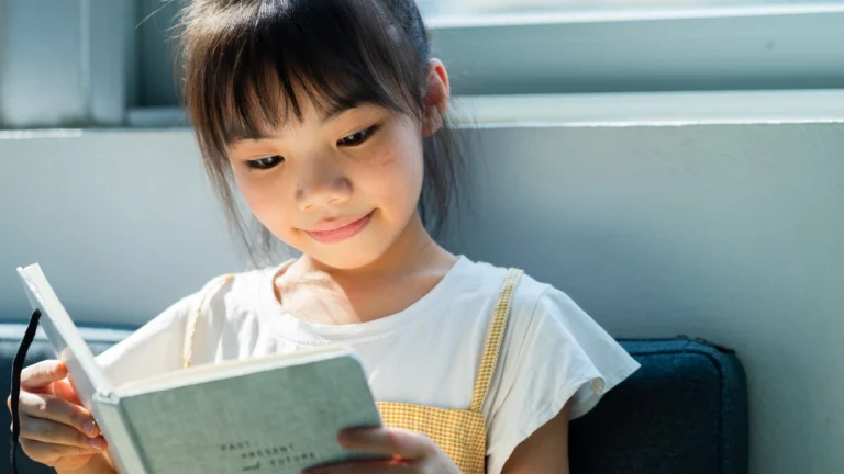A young girl with bangs and a white shirt reads a book while sitting indoors, smiling gently in natural light—a heartwarming moment that captures the joy of teaching reading at home.