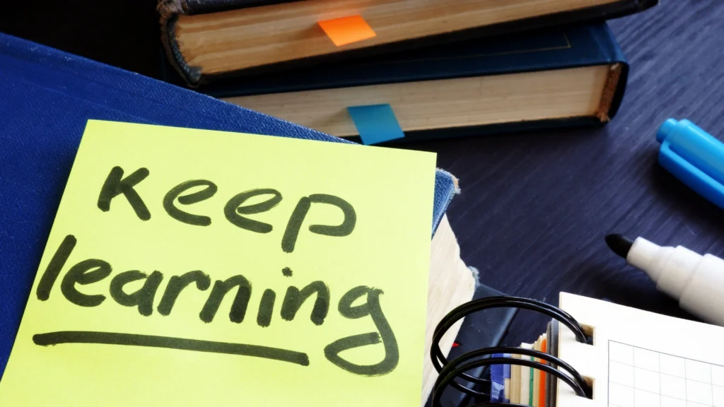 A yellow sticky note with Keep learning written on it sits on top of books with colored bookmarks, embodying a Homeschool Learning Style. Nearby are a marker, blue pen, and notebook with a ring binder.