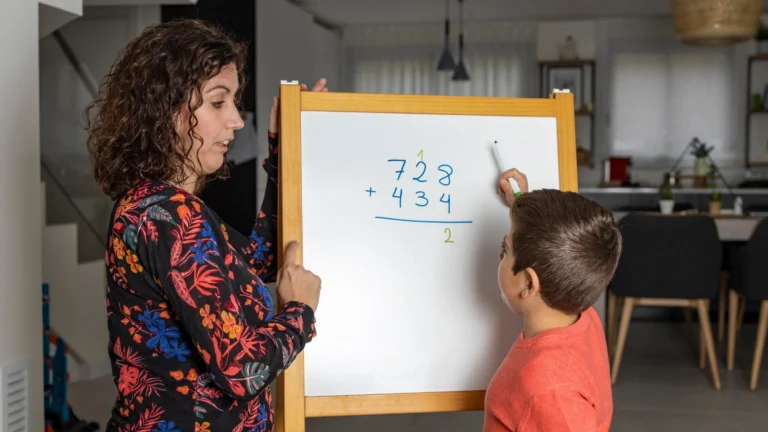 A woman helps a child solve a math addition problem on a whiteboard in a home setting, using their homeschool math curriculum. The child is writing while the woman observes and guides him through each step.