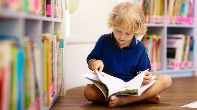 A young child with blonde hair, wearing a blue shirt, sits barefoot on the floor between bookshelves, reading an open book in a library or classroom—a serene moment that captures the joy inspired by a quality Homeschool Reading Curriculum.