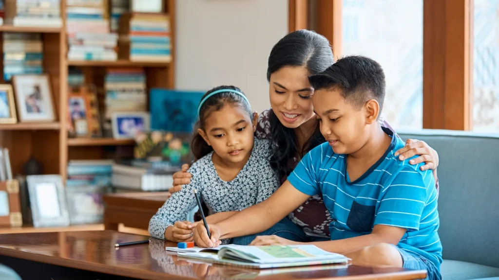 A woman sits with two children at a table, helping them with homework as part of their Homeschool Curriculum Breakdown. The boy writes in a notebook while the girl watches. Bookshelves are visible in the background.