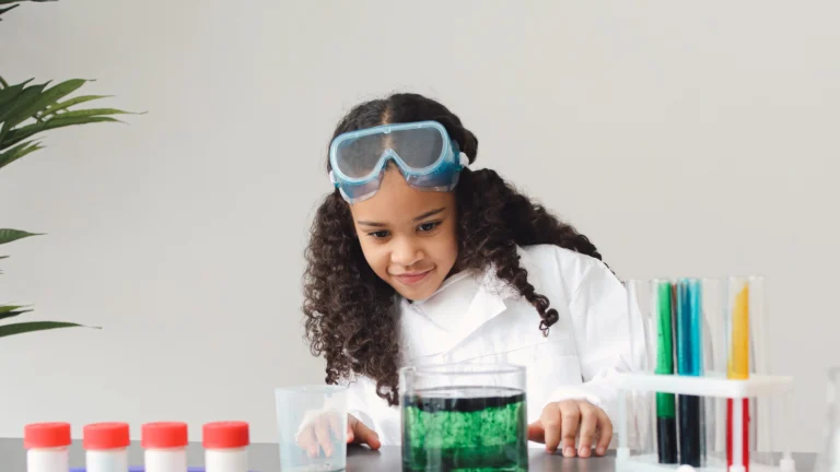 A young girl wearing safety goggles and a lab coat looks curiously at a glass container filled with green liquid, surrounded by colorful test tubes and science equipment on a table—an inspiring scene of hands-on science exploration.