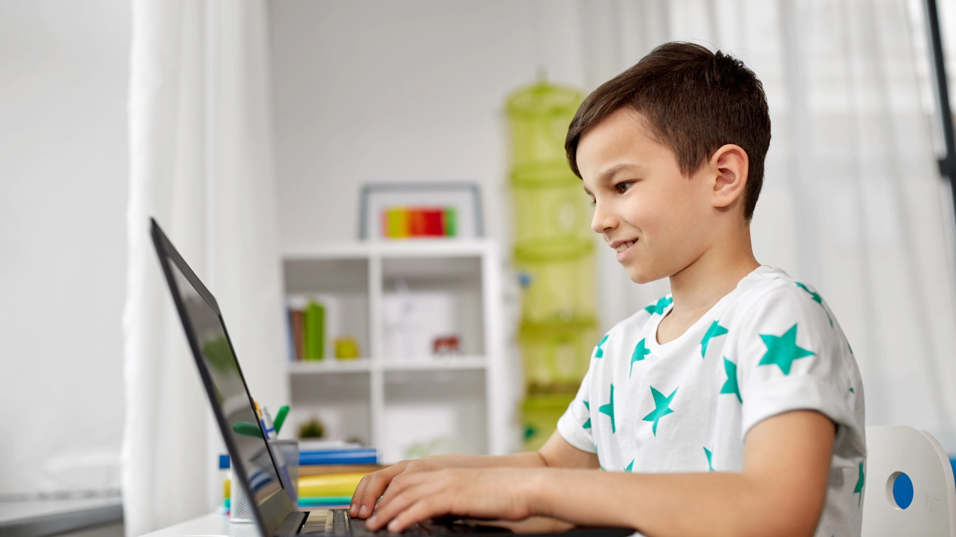 A young boy wearing a white shirt with green stars smiles while using a laptop at a desk in a brightly lit room, exploring some of the Best Online Homeschool Programs, with shelves and books in the background.