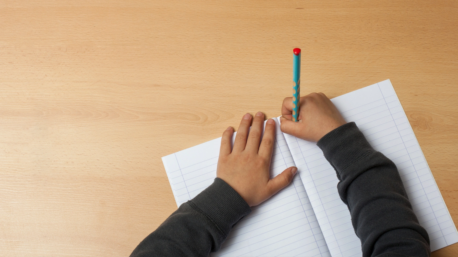 A child wearing a dark long-sleeve shirt writes in a blank, lined notebook with a blue pencil on a light wooden desk. The childs left hand holds the notebook open.