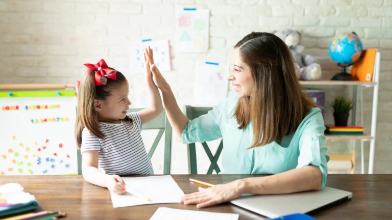 A young girl and an adult woman smile and high-five each other at a table while working on schoolwork, surrounded by papers, pencils, and homeschool resources in the background.