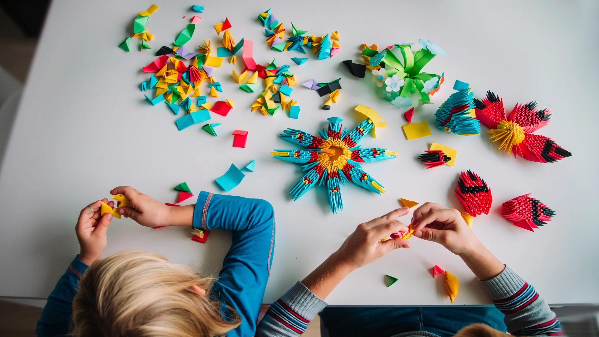 Two children sit at a white table folding colorful origami paper. Several completed origami shapes and scattered paper pieces are visible. The scene is viewed from above.