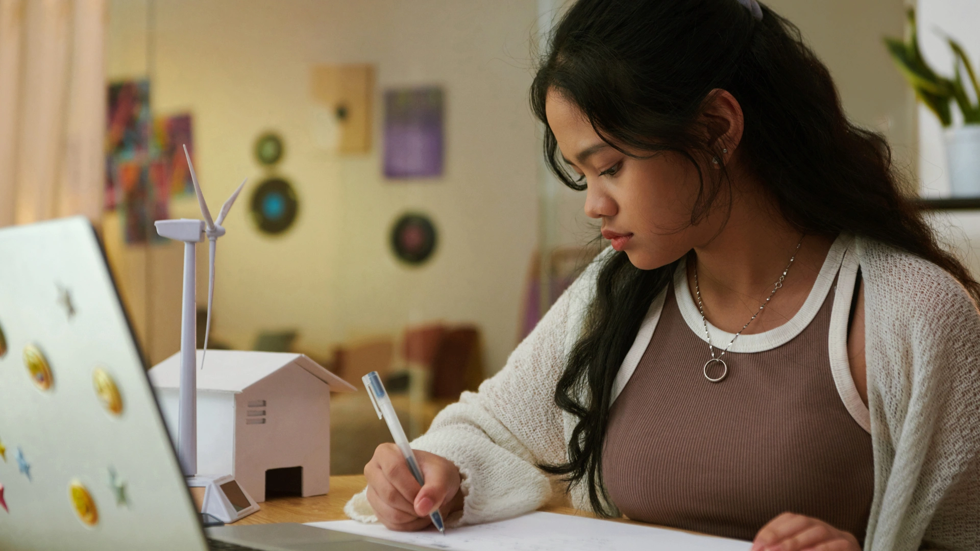 A young woman sits at a desk, writing on paper with a pen. A laptop decorated with stickers and a model house with a wind turbine are in front of her. She appears focused, working in a cozy indoor setting that reflects homeschool gratitude.