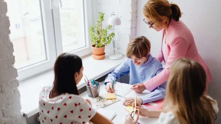 A teacher in a pink blazer helps three children with an arts and crafts project at a table near a window with a potted plant, creating an engaging environment similar to those found in Homeschool Groups Near Me.