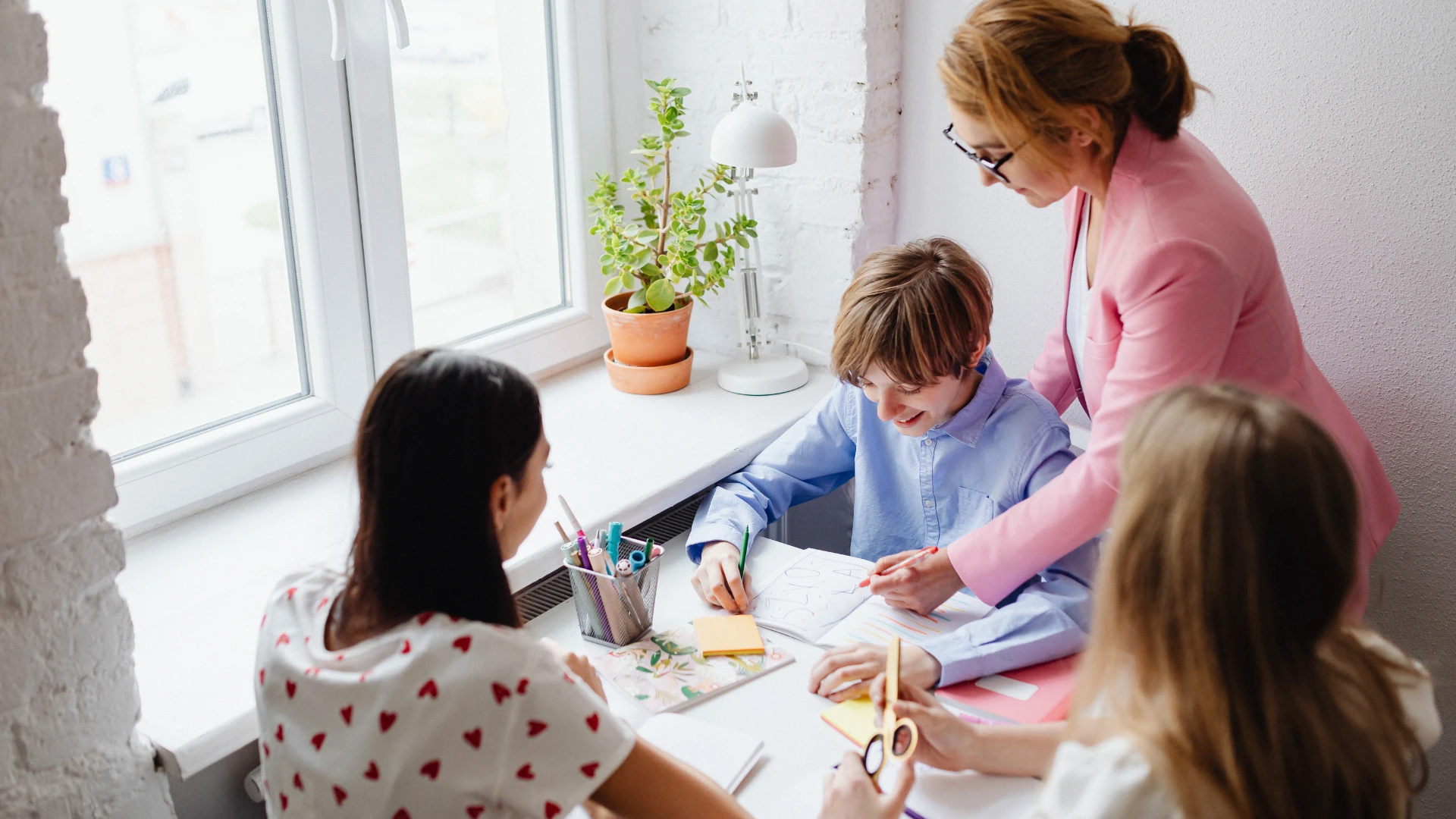 A teacher in a pink blazer helps three children with a creative activity at a table near a window. Perfect for homeschool groups near me, one child draws while others use scissors and colorful paper, surrounded by plants and supplies.