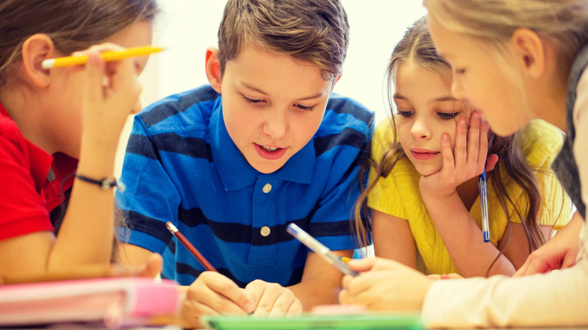 Four children are sitting closely together at a table, writing and drawing on paper with pencils and pens. They appear focused and engaged, fostering reflection and growth as they collaborate in a bright, classroom setting.