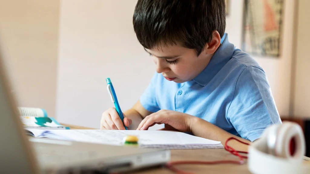 A young boy in a blue shirt sits at a table, focused on writing in a notebook with a blue pen. Beside him, Homeschool Organization Hacks keep his laptop, headphones, and eraser neatly arranged on the desk.