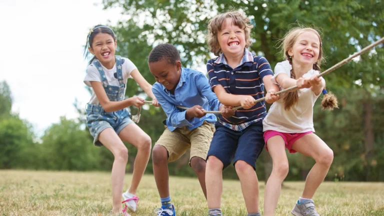 Four smiling children play tug-of-war outdoors on grass, pulling a thick rope together. Trees and greenery are visible in the background, and the kids appear to be having fun on a sunny day, showcasing the benefits of homeschool socialization.