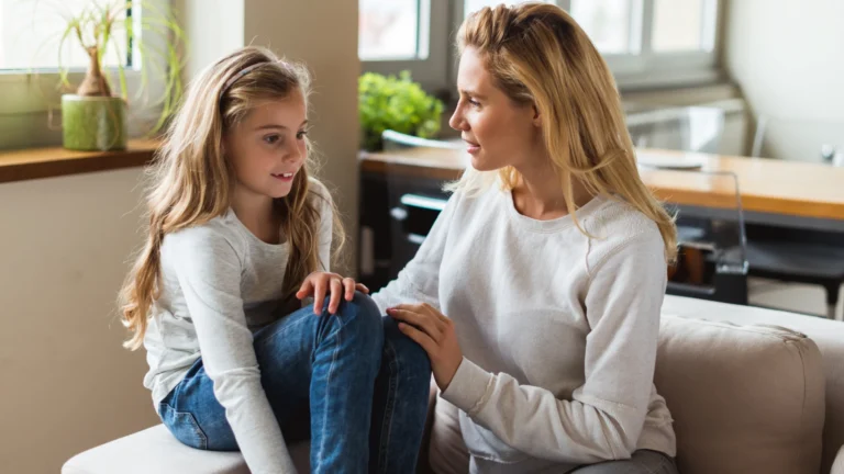 A woman and a young girl sit on a couch indoors, facing each other and talking. Both have long blonde hair and are wearing casual jeans and light sweaters. As natural light fills the room from large windows behind them, they share tips to improve reading fluency in a relaxed, comfortable setting.