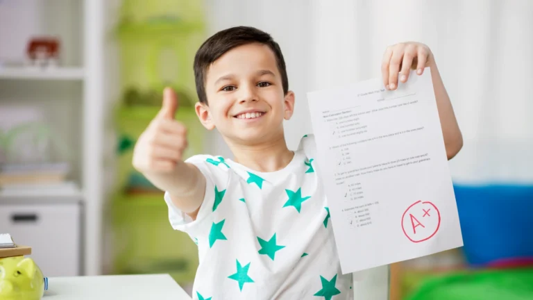 Smiling boy gives a thumbs up while holding a test paper marked with an A+ grade, a perfect way to measure homeschool progress. He wears a white shirt with green stars. The background is brightly lit and out of focus.