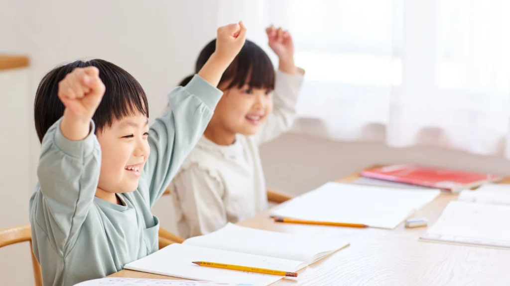 Two young children sit at a desk with open notebooks and pencils, smiling and raising their arms in excitement, likely celebrating a correct answer or achievement in class. Bright, natural light fills the room, creating an atmosphere of joyful learning.