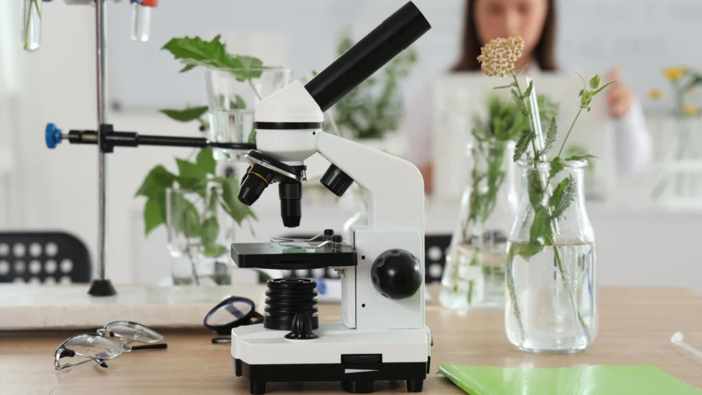 A white microscope sits on a wooden table surrounded by glass vases with green plants, perfect for a Homeschool Science Curriculum Guide. In the background, a person is blurred and out of focus; a green folder and eyeglasses are also on the table.