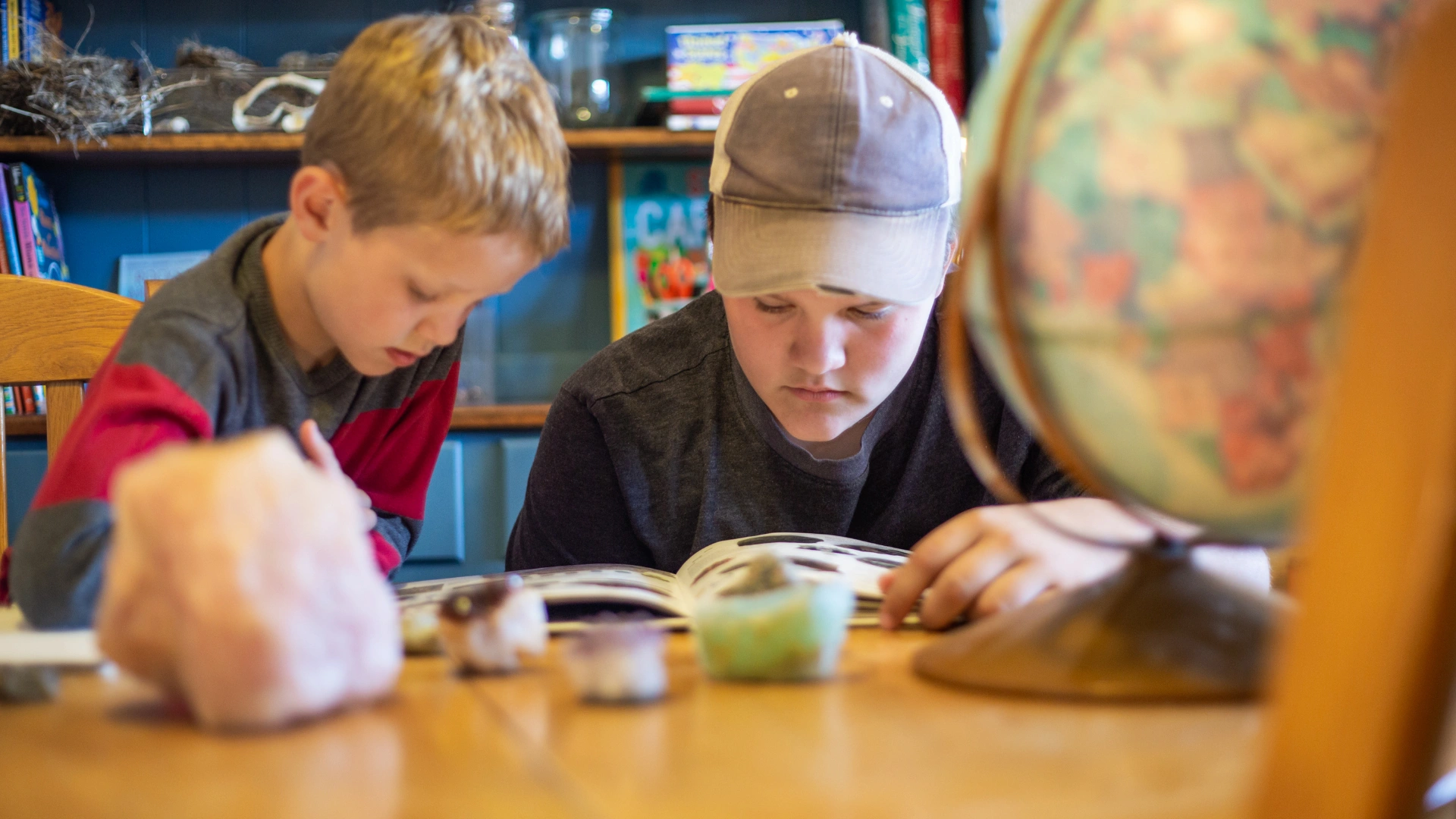 Two boys sit at a table reading a book together, exploring their Homeschool Reading Curriculum, with a globe and various rocks or crystals in the foreground. Bookshelves filled with books are visible in the background.