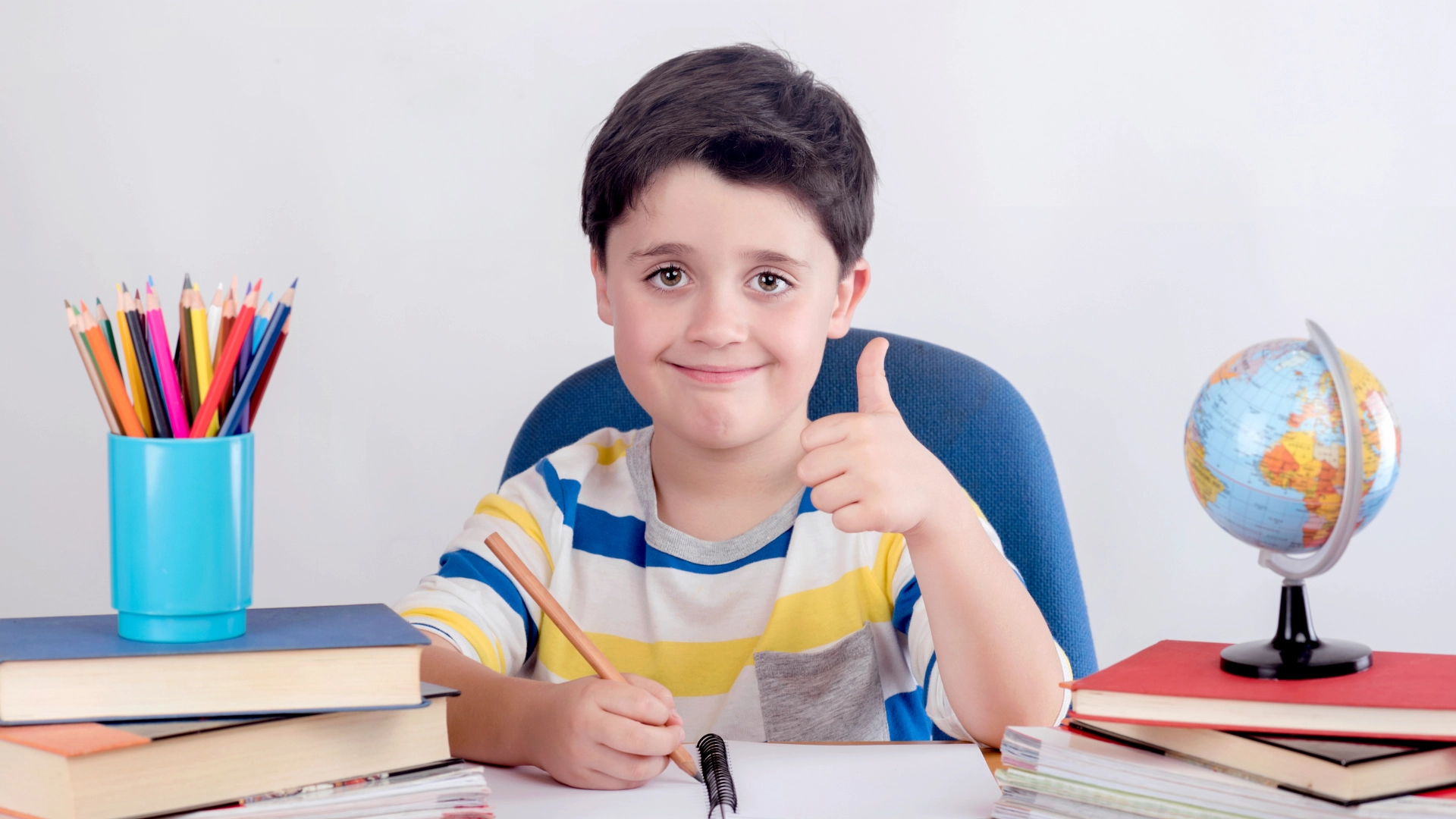 A smiling boy sits at a desk, giving a thumbs up. Surrounded by books, colored pencils, notebooks, and a small globe, he enjoys his Homeschool Social Studies Curriculum in a positive and studious environment.