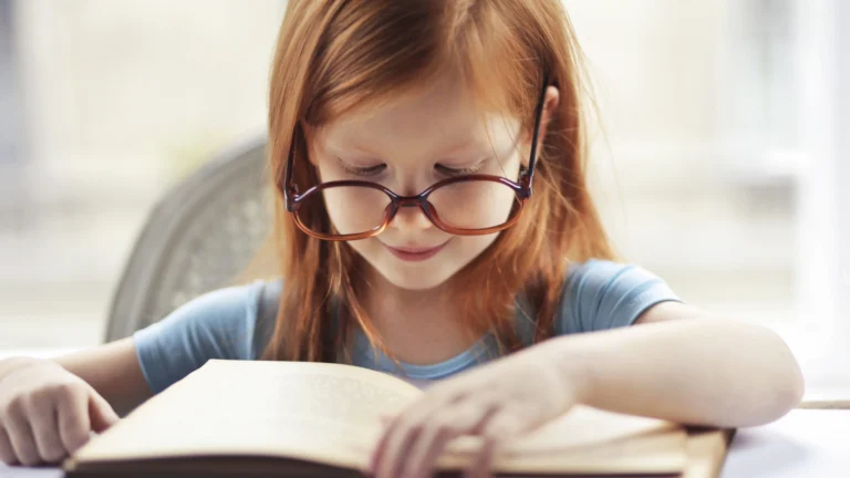 Young girl with long red hair and large glasses reads an open book, concentrating on her reading comprehension while sitting indoors in soft, natural light.